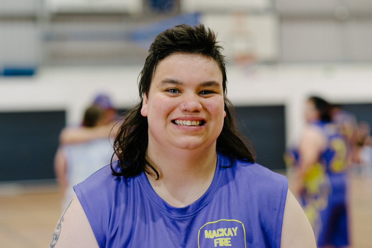 A woman wearing a basketball shirt stands on an indoor court. She is smiling.