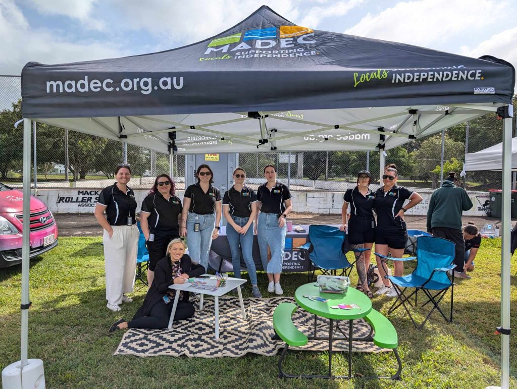 A team of MADEC youth workers stand underneath a MADEC marquee in a park.