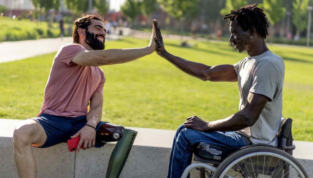 Tow men in a park high five each other. One man has a prosthetic leg and the other is in a wheelchair.